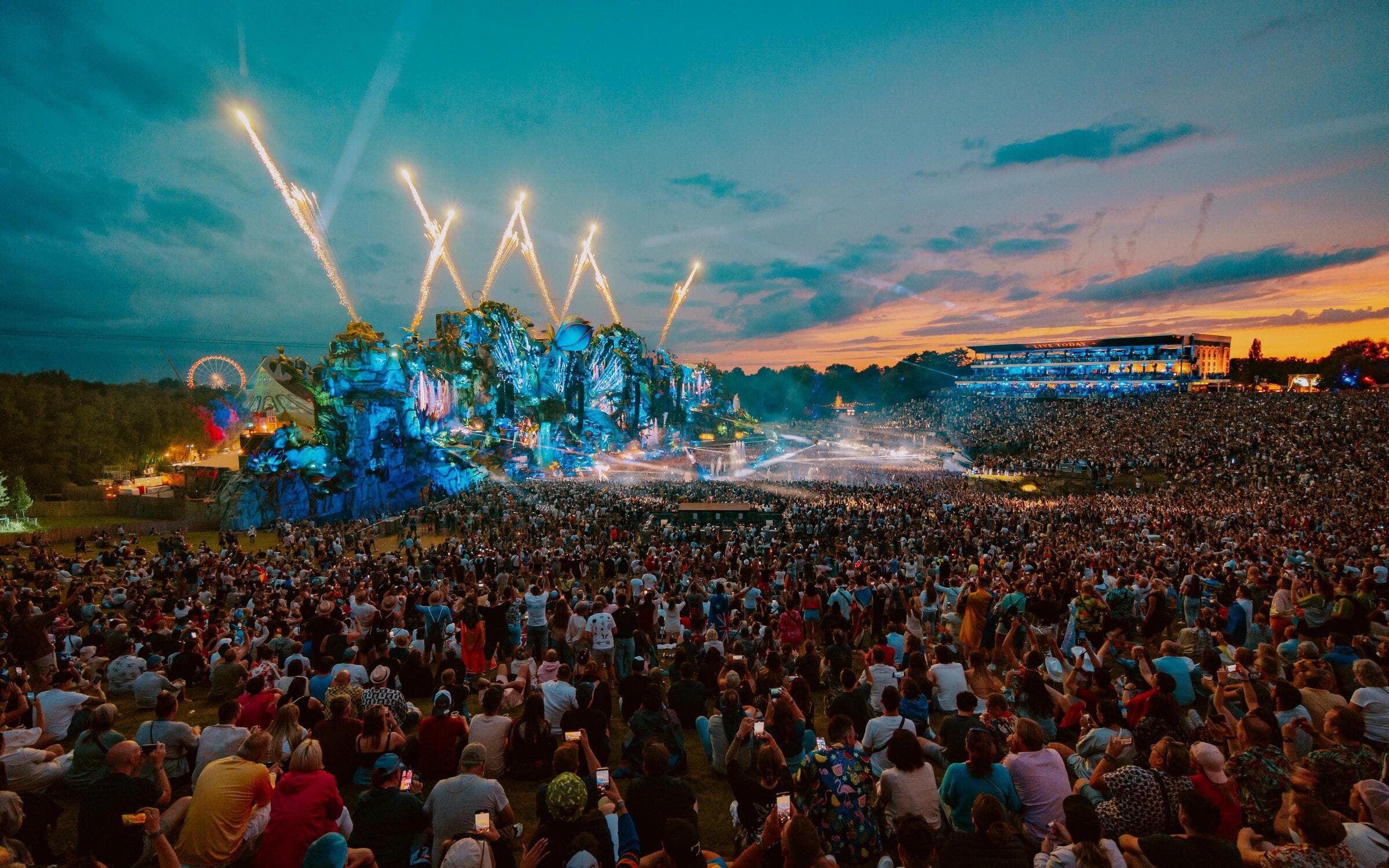 Large crowd watching Tomorrowland’s main stage at sunset during the festival, with fireworks in the sky and thousands of people gathered across the grass field in Boom Belgium.