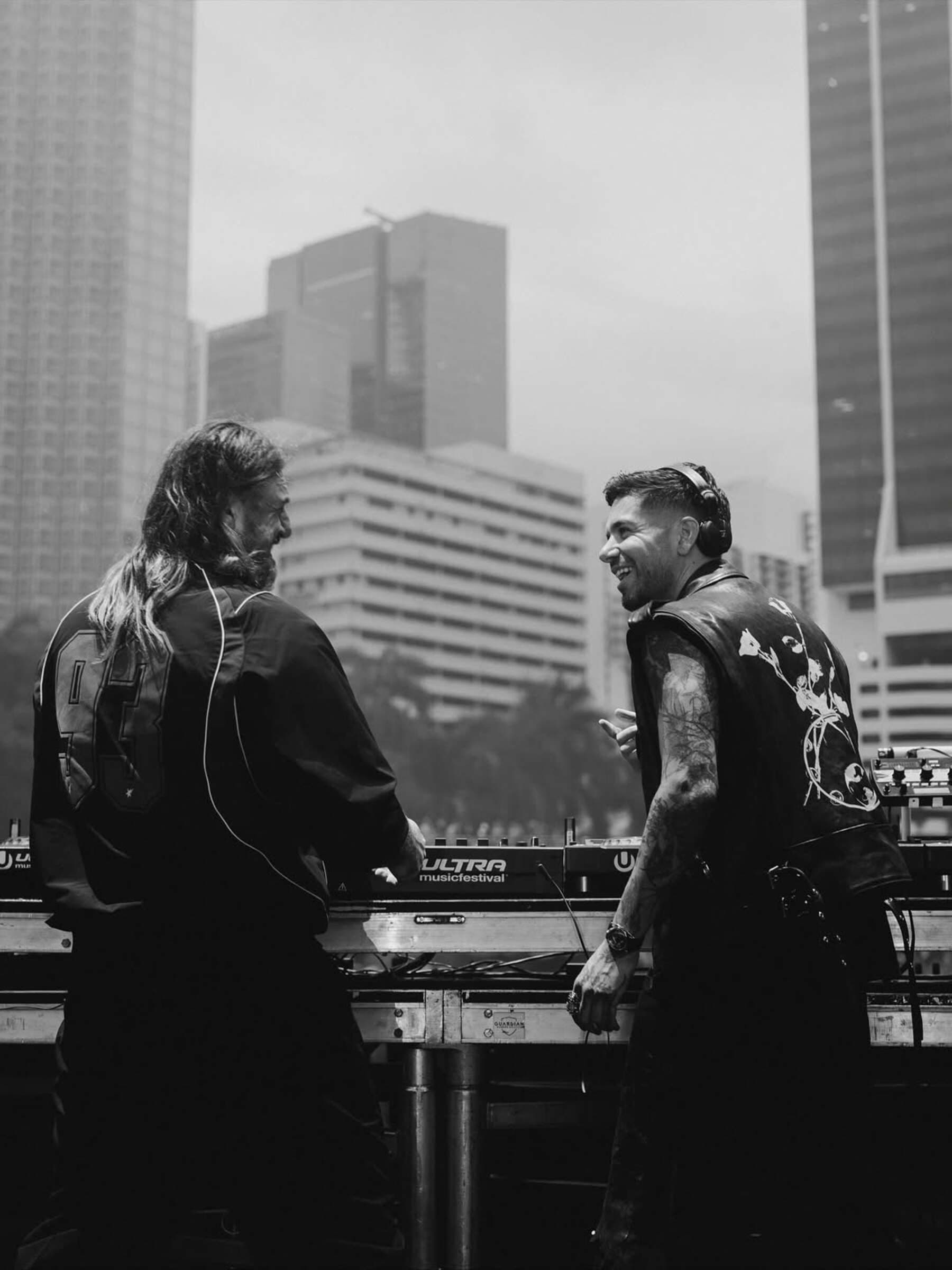 Anyma and Solomun performing together on stage at Ultra Music Festival, smiling and facing each other behind the DJ booth with city skyscrapers in the background.