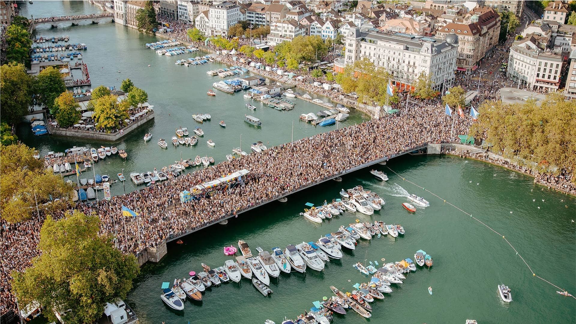 Aerial view of Zurich Street Parade crowds filling the bridge and lakeside, with boats gathered on the water during the 34th edition of the event.