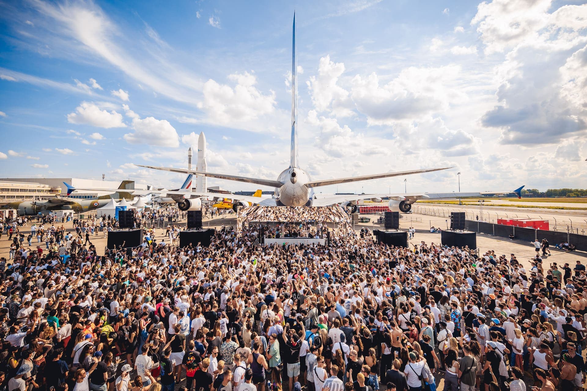 Crowd dancing at Cercle Festival under the wing of a large airplane at the National Air and Space Museum of France, open-air electronic music event at Le Bourget.